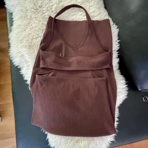 Brun ribbad långklänning med cut-outs - Snygg stretchig brun långklänning i ribbat material med v-ringning och trendiga cut-outs vid midjan. Klänningen är ärmlös och har en tight passform som framhäver figuren. Perfekt för dig som vill sticka ut med en modern och stilren look.