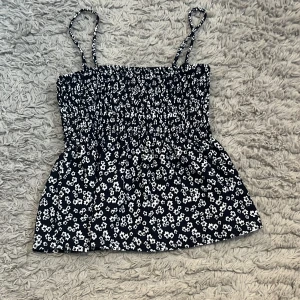 Svart blommig linnetopp med smock - Supersöt svart linnetopp med vita blommor och smockad överdel. Tunt och luftigt material, stretchig passform och smala axelband som går att ta av för en axelbandslös look. Perfekt till jeans eller kjol för en chill vibe.