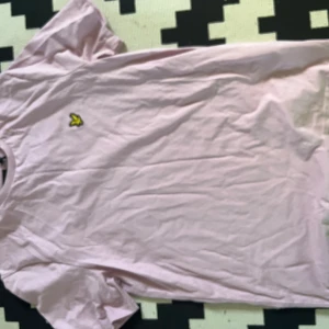 Ljusrosa t-shirt från Lyle & Scott - Snygg ljusrosa t-shirt från Lyle & Scott med klassisk rund hals och korta ärmar. På bröstet sitter den ikoniska gula fågeln som broderad detalj. T-shirten är gjord i mjuk bomull och har en clean, normal passform som funkar till jeans eller shorts.
