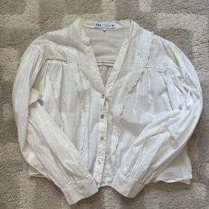 Vit blus med spetsdetaljer från Zara - Supersöt vit blus från Zara med v-ringning, knappar framtill och fina spetsdetaljer längs bröstet. Skulle säga att den är mindre än en m passar S/xs 