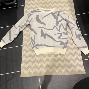 Grå stickad tröja med mönster - Säljer en stickad tröja i ljusgrå och vit med ett abstrakt mönster i mörkare grå toner. Tröjan har rund hals och långa ärmar, perfekt för dig som gillar en chill och stilren look. Passar bra till jeans eller mjukisbyxor.