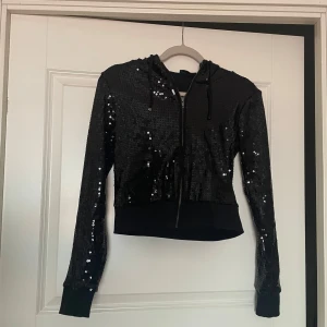 Svart paljett ziphoodie med huva - Snygg svart ziphoodie med dragkedja och huva, täckt av glittriga paljetter. Rätt så Croppad modell med långa ärmar och ribbad längst ner. Perfekt för dig som vill sticka ut och addera lite extra bling till din outfit. Skriv om du har frågor eller vill ha fler bilder, tveka inte🥰
