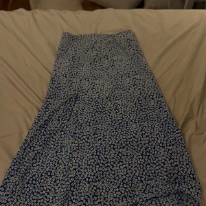 Blommig blå långkjol - Supersöt långkjol med blå botten och små vita blommor över hela tyget. Kjolen har en rak och flowy passform som ger en chill vibe. Perfekt för dig som gillar blommigt och vill ha något som sticker ut lite extra i garderoben.
