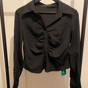 Svart skjorta med rynkade detaljer SHEIN - Svart skjorta från SHEIN i storlek M. Skjortan har långa ärmar, klassisk krage och knappar framtill. Snygg rynkad detalj längs knappraden ger en trendig look. Tillverkad i polyester för en lätt och smidig känsla.