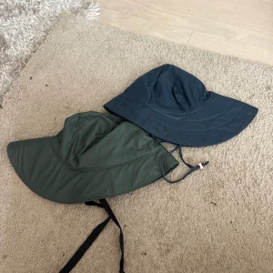 Två bredbrättade hattar med knytband - Säljer två bredbrättade hattar, en i mörkgrönt och en i mörkblått. Båda har knytband under hakan och ljus insida. Perfekta för soliga dagar när du vill ha extra skydd och stil. Materialet är syntetiskt och känns lätt och smidigt.