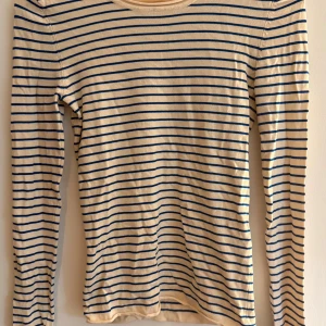 Randig långärmad tröja från Zara - Beige långärmad tröja från Zara med smala blå ränder. Tröjan har rund halsringning och är gjord i mjuk bomull, perfekt för en chill och stilren look. Passformen är tajt och den funkar lika bra till jeans som kjol.