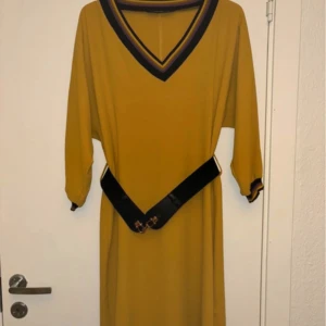 Gul klänning med bälte - Ej använd. Snygg gul klänning med svarta och lila detaljer vid hals och ärmslut. Klänningen har en v-ringad design och ett svart bälte i midjan som ger en fin siluett. Perfekt för en stilren look!