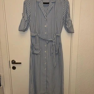 Blåvit randig klänning från Bik Bok - Ej använd bara testad. Snygg blåvit randig klänning från Bik Bok med knappar framtill och ett knytskärp i midjan. Klänningen har trekvartsärmar och en klassisk krage. Perfekt för en stilren look!