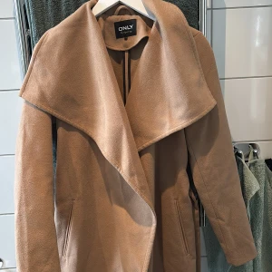 Beige kappa från ONLY - Säljer en stilren beige kappa från ONLY med en elegant draperad krage och bälte i midjan. Perfekt för höst och vår. Kappan har långa ärmar och en mjuk, bekväm känsla.