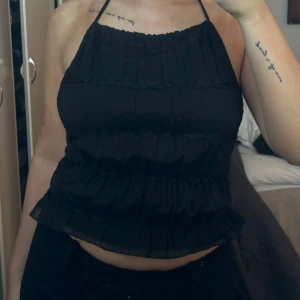 Svart halterneck topp - Snygg svart halterneck topp med volangdetaljer och öppen rygg. Perfekt för en somrig look eller en utekväll. Toppen har knytband i nacken och ryggen för justerbar passform.