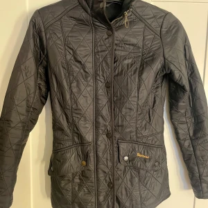 Svart Barbour quiltad jacka  - Snygg svart quiltad jacka från Barbour med dragkedja och knappar. Jackan har en klassisk design med två framfickor och Barbour-logga broderad på ena fickan. Perfekt för kyligare dagar.