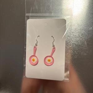 Rosa stekpanna örhängen - Unika örhängen formade som små rosa stekpannor med ett stekt ägg i mitten. Perfekta för att ge en lekfull touch till din outfit. Hängande design med krokfäste. (Billigt material) 