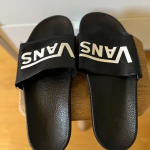 Svarta sandaler från Vans - Säljer ett par svarta sandaler från Vans med en bred rem över foten och logotyp i vitt. Sandalerna har en mönstrad sula för bättre grepp. Perfekta för sommarens varma dagar.