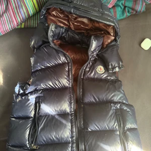 Moncler bormes gilet - Snygg mörkblå dunväst från Moncler med glansig yta och dragkedja framtill. Västen har en hög krage och praktiska fickor med dragkedjor. Perfekt för sommaren när du vill hålla stilen. Kvitto finns, pris ej hugget i sten. 