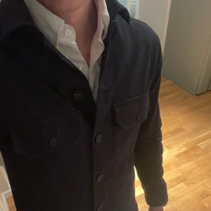 Mörkblå overshirt från The Shirt Factory - Snygg mörkblå overshirt från The Shirt Factory med knappar framtill och två bröstfickor. Perfekt för lager-på-lager-stil och passar till många olika outfits. En stilren och mångsidig jacka perfekt för vår och sommarkvällar! (Pris kan diskuteras)