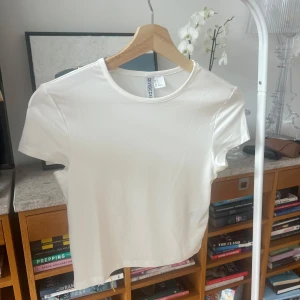 Vit kortärmad topp - Säljer en stilren vit kortärmad topp från H&M Divided. Toppen är gjord i ett mjukt och stretchigt material av polyester och elastan, vilket ger en skön passform. Perfekt för en enkel och fräsch look. Cropped.