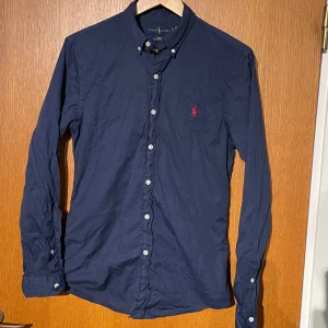 Mörkblå skjorta från Ralph Lauren - Säljer denna snygga mörkblå skjortan från Ralph Lauren i slim fit. Stl S. Skjortan har långa ärmar och knappar framtill. Den har en röd broderad raffe logga på bröstet. Säljer för att den inte kommer till användning. Skjortan är lite skrynklig på bilderna men stryker självklart till den innan jag skickar. Kom gärna med frågor.