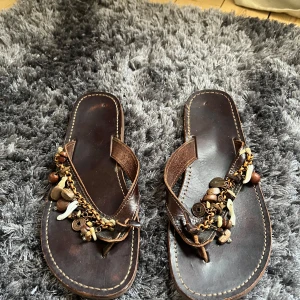 Bruna sandaler med dekorativa detaljer - Säljer ett par bruna sandaler med dekorativa kedjor och pärlor. Sandalerna har en platt sula och är perfekta för sommarens varma dagar. De har en avslappnad och bohemisk stil som passar bra till många olika outfits.