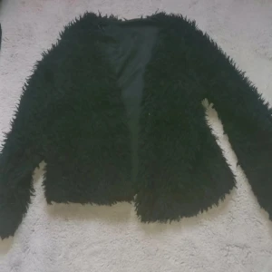 Svart pälskofta - Säljer en svart pälskofta med långärmad design. Koftan har en fluffig textur och är perfekt för att hålla värmen eller vill fixa till sig lite extra.. Den har en öppen framsida utan knappar eller dragkedja.