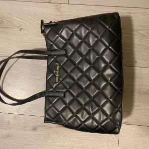 Svart quiltad axelväska  - Snygg svart axelväska från Valentino i quiltat mönster. Väskan har två långa handtag och en dragkedja upptill. Perfekt för att bära med sig alla nödvändigheter med stil.