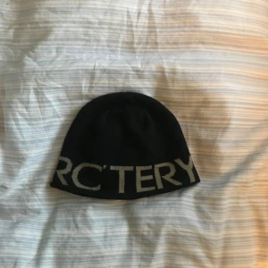 Svart mössa från Arc'teryx - Snygg svart mössa från Arc'teryx med grå textdetalj. Perfekt för kyliga dagar och en stilren look. Passar både till vardags och utomhusaktiviteter.