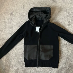 Cardigan med huva från Roy Robson - Snygg svart jacka från Roy Robson med huva och dragkedja. Jackan har två stora fickor framtill med kontrasterande material. Perfekt för en stilren look. Jackan är i Size M och har en otrolig passform, jackan är helt ny med lappen kvar samt så har den en otrolig kvalite, kom dm för frågor! 