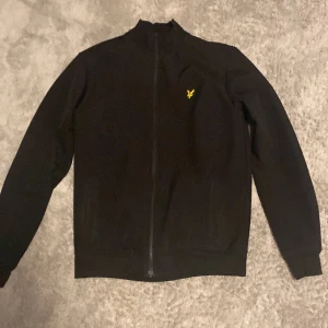 Svart jacka med dragkedja och broderad logga - Snygg svart jacka med dragkedja och en gul broderad logga på bröstet. Jackan har långa ärmar och en stilren design, perfekt för en casual look. Nypris 900kr. Priset är ej hugget i sten