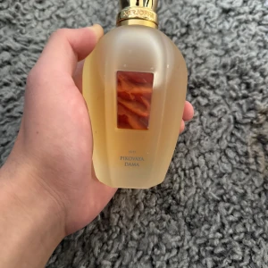 Parfym Firovaya Dama - Elegant parfymflaska med en frostad yta och gyllene lock. Flaskan har en rektangulär röd detalj i mitten som ger en lyxig känsla. Perfekt för den som söker en unik doftupplevelse.