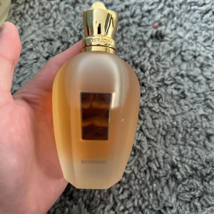 Richwood Parfym från Xerjoff - Elegant parfymflaska från Xerjoff med namnet Richwood. Flaskan har en frostad yta med en gyllene kork och detaljer. Den har en rektangulär etikett på framsidan. Perfekt för den som söker en lyxig doftupplevelse.