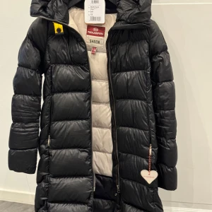 Svart dunjacka från Parajumpers - Snygg svart dunjacka från Parajumpers med dragkedja och lång passform. Jackan har en hög krage och är perfekt för kyliga dagar. Använd endast en säsong. 