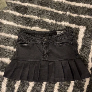 Svart jeanskjol med volang - Snygg svart jeanskjol med volangdetalj. Kjolen har en klassisk femficksdesign och en kort längd som ger en trendig look. Perfekt för att ge din outfit en cool touch.