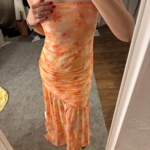 Orange axelbandslös klänning från ADOORE - Säljer en snygg axelbandslös klänning från ADOORE i en härlig orange färg med mönster. Klänningen har en smickrande rynkad design och en volangdetalj vid fållen. Perfekt för sommarens fester!Storlek 42(liten i storleken) 