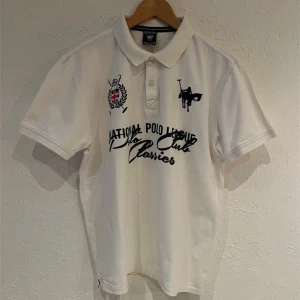 Vinson Polo Club Vit pikétröja - Strl L - Snygg vit pikétröja från Vinson Polo Club med broderade detaljer och text på framsidan. Tröjan har en klassisk krage och knappar framtill. Perfekt för en stilren look!                                                                                    100% Bomull