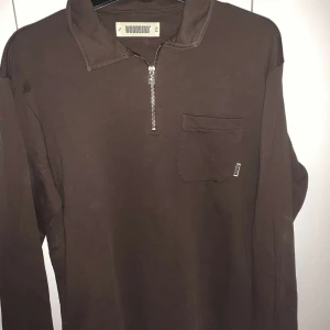 Brun tröja med dragkedja från Woodbird - Säljer en brun tröja från Woodbird med dragkedja och bröstficka. Tröjan har långa ärmar och en stilren design, perfekt för en avslappnad look.