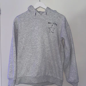 Grå hoodie från Gina Tricot - Säljer en grå hoodie från Gina Tricot med texten 'MA CHÉRIE' och en stjärna tryckt på både fram- och baksidan. Tröjan har en klassisk huva med dragsko och långa ärmar. Perfekt för en avslappnad stil.