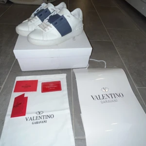 Valentino open - Snygga vita Valentino Garavani sneakers med en bred mörkblå rem över snörningen. Skorna har en klassisk design med rund tå och är tillverkade i skinn. Perfekta för en stilren look. På ens skon är det bytt snöre över de mörkblåa. Storlek 43, finns något skav på skon men kom privat för bild