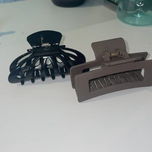 Hårklämmor i svart och beige - Två stilrena hårklämmor i svart och beige. Den svarta klämman har en klassisk design med rundade kanter, medan den beige klämman har en modern, rektangulär form. Perfekta för att hålla håret på plats med stil.