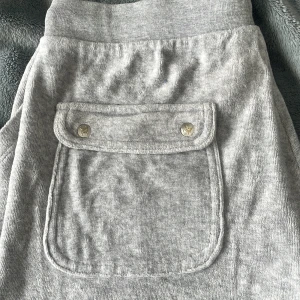 Grå mjukisbyxor från Juicy Couture - Säljer ett par grå mjukisbyxor från Juicy Couture. Byxorna har en praktisk ficka med knappar baktill och snörning i midjan för justerbar passform. Perfekta för en avslappnad stil. De säljs pga att de är för stora och används ej längre. Skriv till mig innan du köper!!
