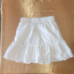 Vit kjol med volang - Söt vit kjol med volangdetaljer och elastisk midja. Perfekt för en somrig look med sitt lätta och luftiga material. Kjolen har ett diskret broderat mönster som ger en extra touch av stil.