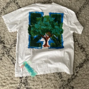 Vit t-shirt Off-White - Säljer en vit t-shirt från Off-White med ett stort färgglatt trädtryck på baksidan. Framsidan har en minimalistisk design med små tryck. Perfekt för dig som gillar unika och konstnärliga plagg.