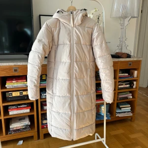 Beige lång pufferjacka från Ellos - Säljer en stilren beige lång pufferjacka från Ellos. Jackan har en praktisk dragkedja framtill och en mysig huva. Perfekt för kyliga dagar med sin långa design och quiltade mönster.
