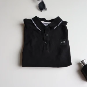 Boss piké  - Säljer en  svart pikétröja fån boss med vita detaljer på kragen. Tröjan har korta ärmar och en klassisk pikéknäppning framtill. 