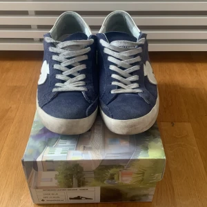 Blå sneakers från The Gallerist - Snygga blå sneakers från The Gallerist med vita detaljer och snörning i storlek 43. Skorna har gått upp lite längst bak på skorna men är enkelt att fixa och syns knappt. Skorna är perfekta för sommaren och våren.