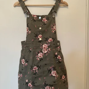 Blommig hängselklänning - Säljer en snygg hängselklänning från H&M i en härlig grön färg med rosa blommönster. Klänningen har justerbara axelband och knappar framtill. Perfekt för sommaren.