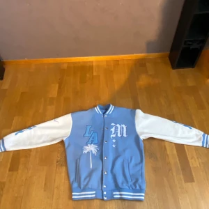 Blå och vit bomberjacka med broderier - Snygg bomberjacka i blått och vitt med broderade detaljer som 'LA' och palmer. Jackan har långa ärmar och knappar framtill. Perfekt för en cool och avslappnad stil.