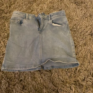 Blå jeanskjol från Your Denim - Säljer en snygg blå jeanskjol från Your Denim. Kjolen har en klassisk femficksdesign med knapp och dragkedja framtill. Perfekt för en avslappnad stil. Passar storlek 134/140.