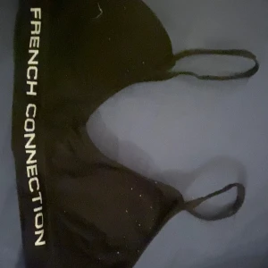 Svart linnetopp från French Connection - Snygg svart linnetopp från French Connection med smala axelband och logga längs sidan. Perfekt för en stilren look. Tillverkad i en mjuk och stretchig nylonblandning.