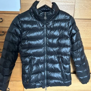 Svart dunjacka från Moncler - Snygg svart dunjacka från Moncler med dragkedja och hög krage. Jackan har långa ärmar och två fickor med dragkedja framtill. Perfekt för kyligare dagar.