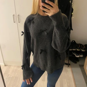 Mörkgrå tröja med rosetter - Säljer en mysig mörkgrå tröja dekorerad med svarta rosetter. Tröjan har långa ärmar och en avslappnad passform, perfekt för kyliga dagar. Den är både stilren och unik med sina fina detaljer. Perfekt för dig som vill ha något extra i garderoben!