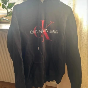 Svart hoodie från Calvin Klein Jeans - Säljer en svart hoodie från Calvin Klein Jeans med deras ikoniska logga i rött och vitt på framsidan. Tröjan har en klassisk känguruficka och justerbar huva med dragsko. Perfekt för en avslappnad stil.
